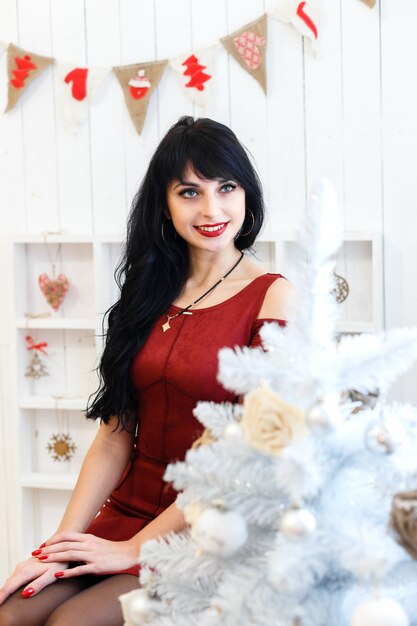 La muchacha triguena joven feliz en el vestido rojo que se sentaba dentro en Navidad adornó el interior.