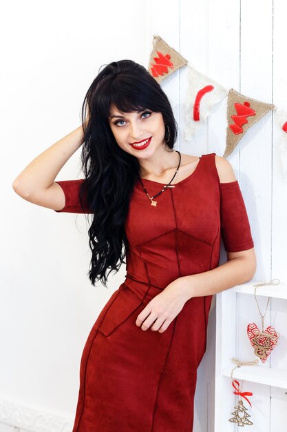 La muchacha triguena joven hermosa en vestido rojo sitanding en una Navidad adornó el interior.