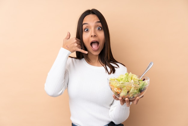 Muchacha triguena joven que sostiene una ensalada sobre la pared aislada que hace gesto del teléfono. Llámame señal de vuelta