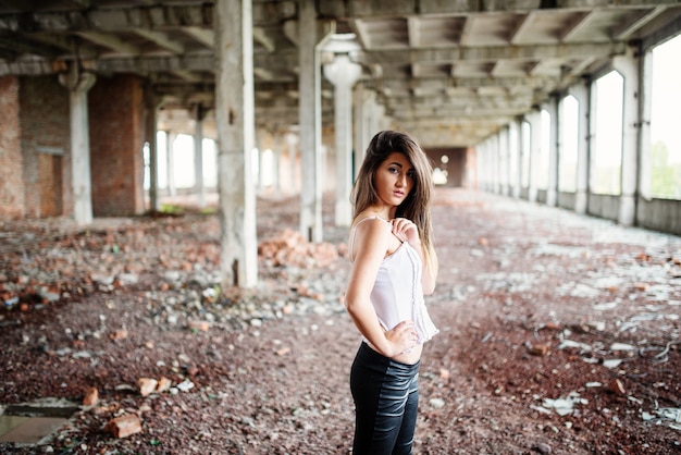 La muchacha triguena linda joven que llevaba en los pantalones de cuero negros y la blusa blanca presentó en lugar abandonado.