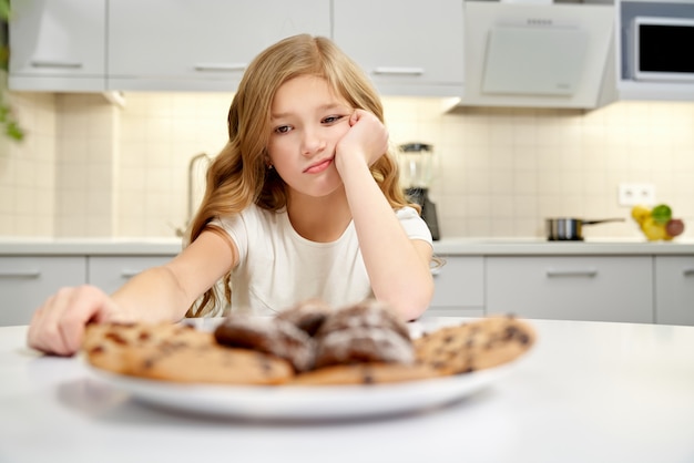 Muchacha triste que mira las galletas americanas, sentándose en cocina.