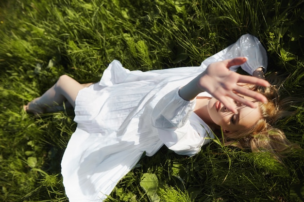 La muchacha en un vestido blanco largo miente en la hierba en un campo. Mujer rubia en el sol en un vestido ligero. Chica descansando y soñando, maquillaje de verano perfecto en su rostro
