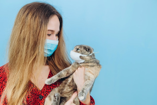 La muchacha en un vestido rojo con una máscara médica en su cara sostiene un gato.