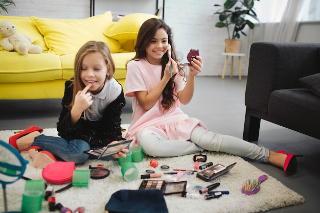 Las muchachas alegres y emocionadas se sentaron en piso en sitio. Ellos haciendo maquillaje. Los adolescentes se miran en el espejo y usan cosméticos.