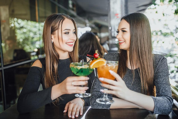 Las muchachas hermosas jóvenes están sentadas en la terraza de verano de un café moderno y beben cócteles refrescantes en naranja y verde.