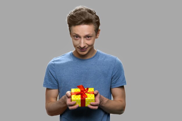 Muchacho adolescente atractivo que da la caja actual. Chico guapo adolescente con caja de regalo sobre fondo gris. Feliz día de la madre.