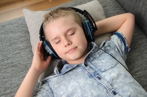 Muchacho adolescente en auriculares escucha música en el sofá en casa. Adolescente disfruta de la música - retrato de niño rubio mientras está acostado en el sofá en el interior. Tiempo de relajación.