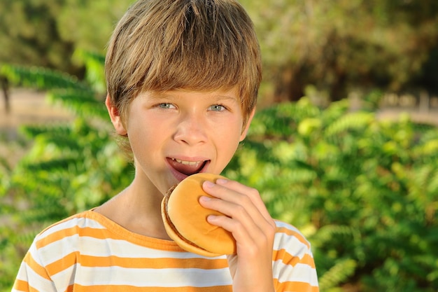 Muchacho adolescente feliz comiendo hamburguesas al aire libre sobre fondo de naturaleza. Comida chatarra poco saludable