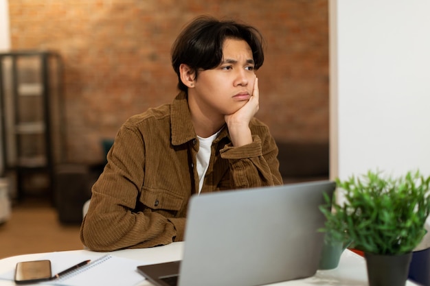 Muchacho adolescente japonés deprimido sentado en la computadora portátil en casa