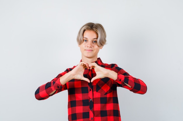 Muchacho adolescente joven en camisa a cuadros que muestra el gesto del corazón y que mira amable, vista frontal.