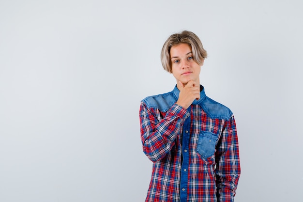 Muchacho adolescente joven que mantiene la mano en la barbilla en la camisa a cuadros y que parece sensible, vista frontal.