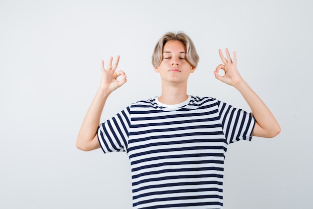 Muchacho adolescente joven que muestra el signo de mudra, manteniendo los ojos cerrados en una camiseta a rayas y mirando relajado, vista frontal.