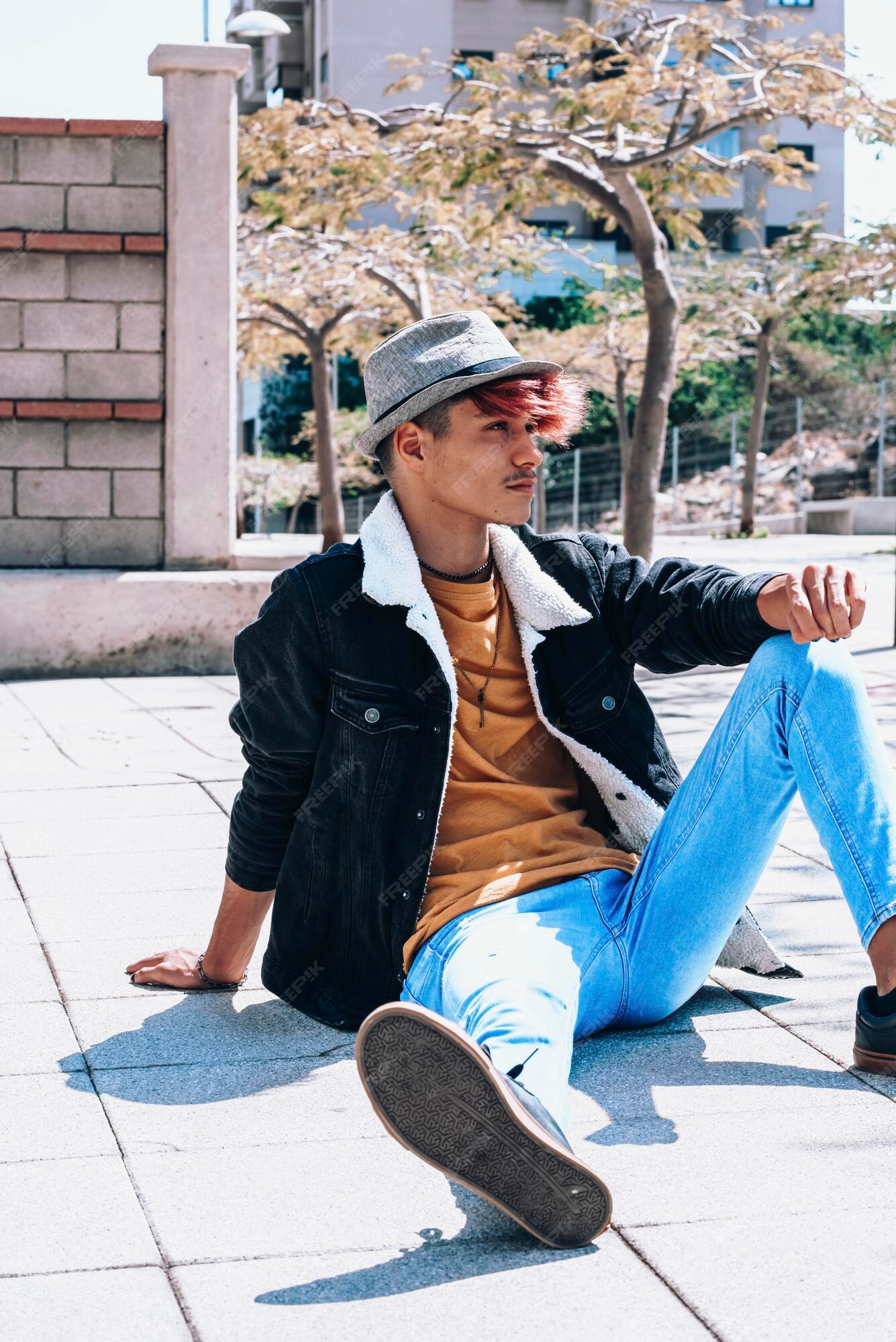 Muchacho adolescente con ropa de estilo urbano sentado el suelo de asfalto en la ciudad chico moderno con sombrero pose de aspecto alternativo en el centro de la ciudad los