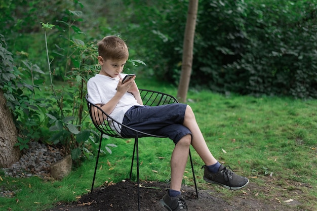 Muchacho adolescente se sienta en una silla en la naturaleza y mirando el teléfono. Gadgets del hombre moderno