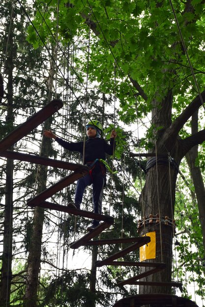 Un muchacho adolescente sube las escaleras colgantes de un árbol a otro en un parque de atracciones de cuerda