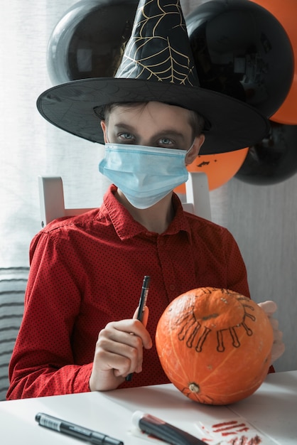 Muchacho adolescente en traje que se prepara para la celebración de Halloween dibujando una calabaza. Carnaval de Halloween con nueva realidad con concepto pandémico