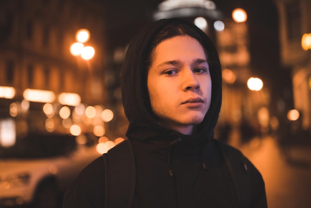 Muchacho adolescente usa chaqueta negra y sudadera con capucha de pie en la calle sobre las luces brillantes de la ciudad