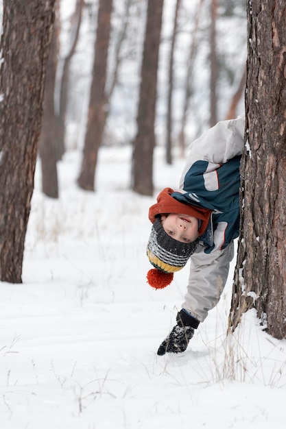 Muchacho alegre se asoma detrás de un árbol. Niño con gorro de punto camina por el parque cubierto de nieve de invierno. Marco vertical