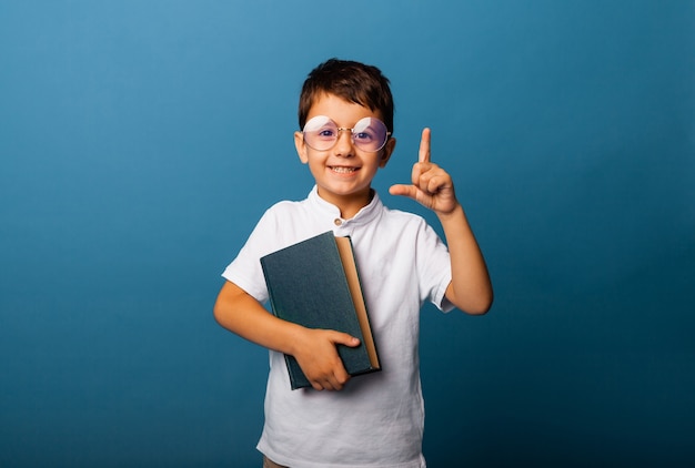 Muchacho alegre, caucásico, sosteniendo un libro en sus manos. niño con un libro señala con el dedo hacia arriba sobre un fondo azul.