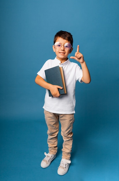 Muchacho alegre, caucásico, sosteniendo un libro en sus manos. niño con un libro señala con el dedo hacia arriba sobre un fondo azul.