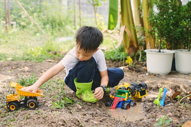 Muchacho asiático con botas jugando en charcos fangosos cavando en suelo fangoso con camión de juguete en casa