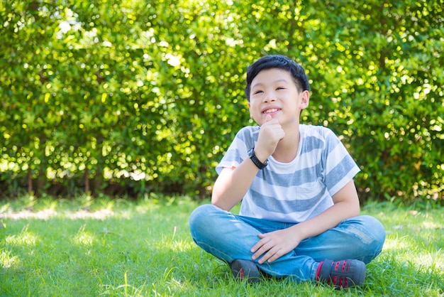 El muchacho asiático joven que se sienta en jardín y piensa con sonrisas