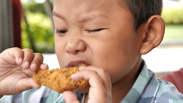 El muchacho asiático lindo es feliz que come la pierna de pollo frito en restaurante.