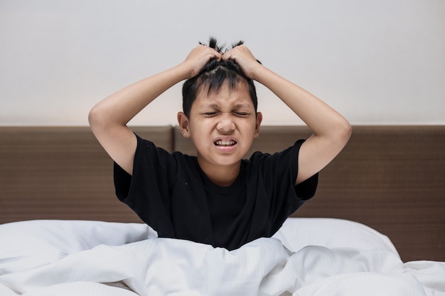 Muchacho asiático sentado en la cama sosteniendo su cabeza sufriendo dolor de cabeza