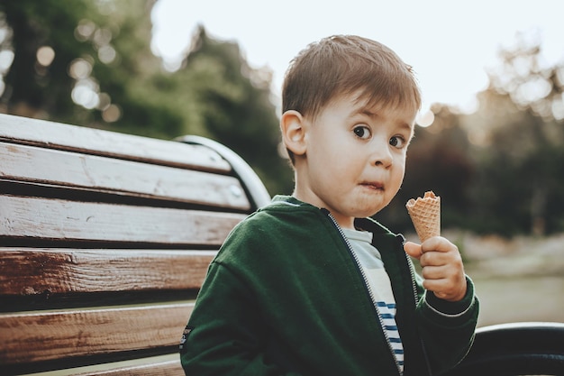 Muchacho caucásico comiendo helado y mirando sorprendido a la cámara mientras está sentado en el banco en el parque ch...