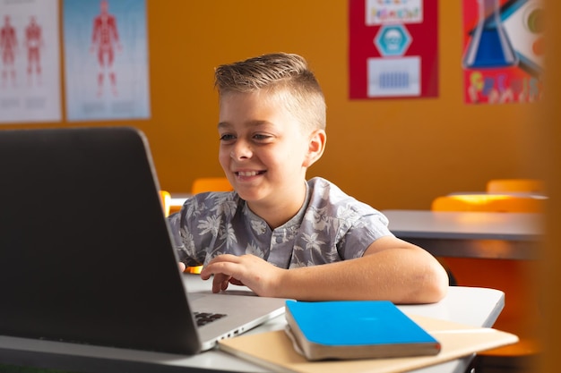 Muchacho caucásico sentado en un escritorio en el aula usando una computadora portátil y sonriendo