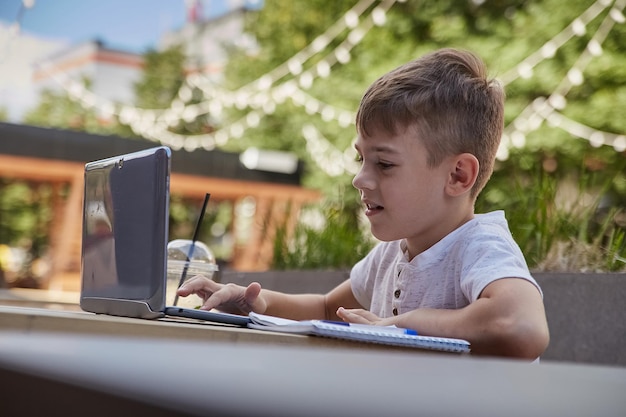 Muchacho caucásico sonriente sentado en la mesa en la veranda del café estudiando con la computadora portátil