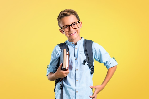 Muchacho del estudiante con la mochila y los vidrios que presentan con los brazos en la cadera en fondo amarillo.
