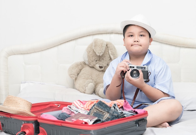 Muchacho gordo feliz que sostiene la cámara vintge. Después de los trajes de accesorios de viaje preparados en maleta para el