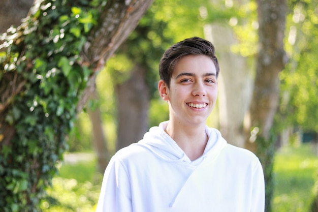 Muchacho hermoso sonriente feliz del adolescente en la sudadera con capucha blanca en parque al aire libre en el día soleado brillante