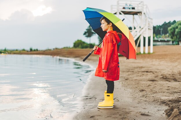 Muchacho con un impermeable rojo y botas de goma amarillas se encuentra en la orilla del río y sostiene el paraguas del arco iris