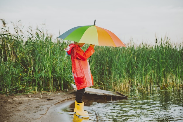 Muchacho con un impermeable rojo y botas de goma amarillas se encuentra en la orilla del río y sostiene el paraguas del arco iris