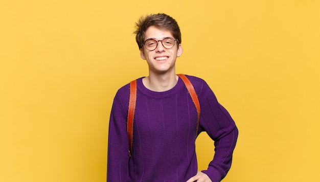 Muchacho joven estudiante sonriendo felizmente con una mano en la cadera y actitud positiva, orgullosa y amistosa confiada