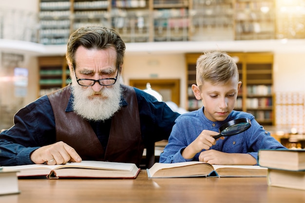 Muchacho lindo que sostiene el libro de lectura de la lupa con su abuelo hermoso, sentado en la mesa en la biblioteca con estilo antiguo, en el fondo de los estantes de libros vintage. Concepto del día mundial del libro