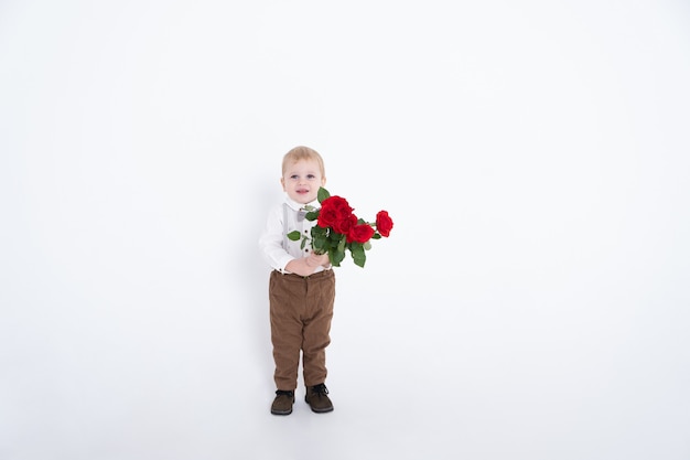 Muchacho del niño del bebé que sostiene el ramo de rosas rojas que sonríen en traje en blanco.
