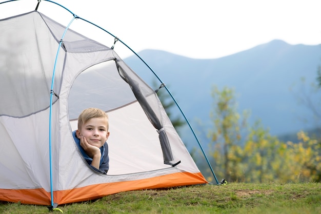 Muchacho del niño excursionista descansando en una tienda turística en el camping de montaña disfrutando de la vista de la hermosa naturaleza de verano.