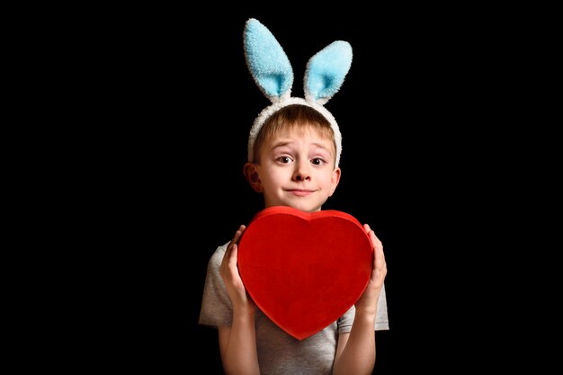 El muchacho rubio divertido en oídos de las liebres sostiene la caja roja de la forma del corazón en fondo negro. Concepto de amor y familia. Copiar spase