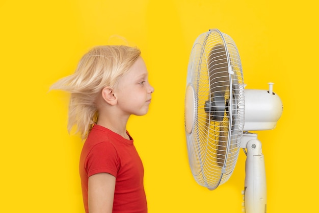 El muchacho rubio está parado cerca del ventilador sobre fondo amarillo brillante. Verano caluroso.