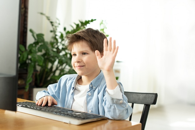 Muchacho sonriente chateando en línea y saludando a la pantalla de la computadora. Niños y artilugios. Aprendizaje a distancia durante el aislamiento durante la cuarentena. Niño y portátil en casa. Estilo de vida