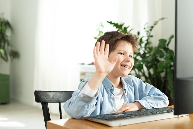 Muchacho sonriente chateando en línea y saludando a la pantalla de la computadora. Niños y artilugios. Aprendizaje a distancia Niño y portátil en casa. Estilo de vida