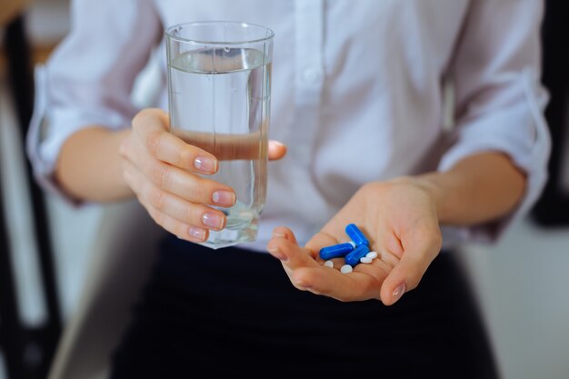 Muchas pastillas. Mujer joven vestida con camisa blanca y sosteniendo un vaso con agua en la mano derecha mientras te va a ayudar