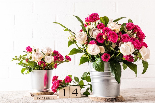 Muchas rosas blancas y rojas en dos cubos decorativos de metal sobre una mesa de mármol contra un calendario de madera de pared de ladrillo blanco con un día de fiesta de San Valentín