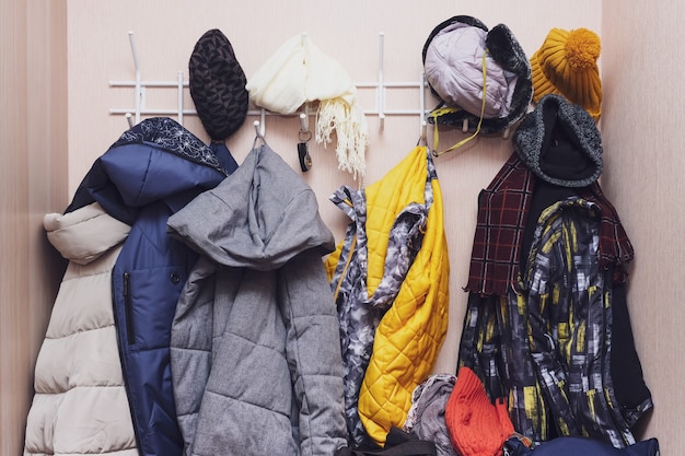 Foto muchos abrigos y gorras de invierno, sombreros colgados desordenados de ganchos en un pasillo, ordenando ropa abrigada.