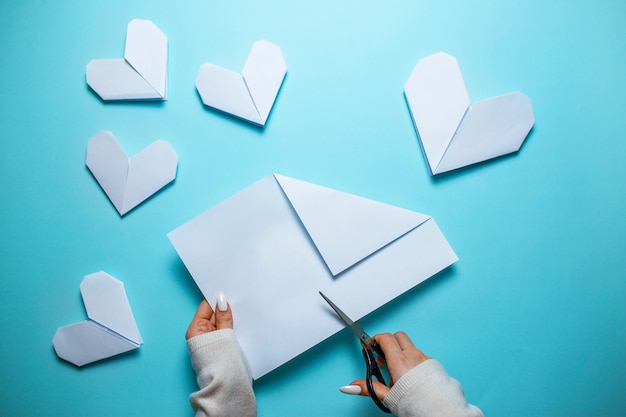 Muchos corazones de origami blanco sobre fondo azul con hoja de papel y tijeras en el centro. Tarjeta del día de San Valentín con mujer haciendo origami corazón sobre fondo azul.