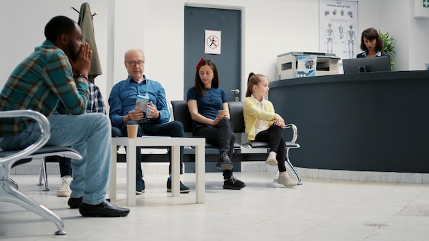 Muchos pacientes sentados en el vestíbulo de la sala de espera en la recepción del hospital, preparándose para comenzar la visita de control con cita. Madre con hijo, anciano y otras personas en la sala de espera. Disparo de trípode.