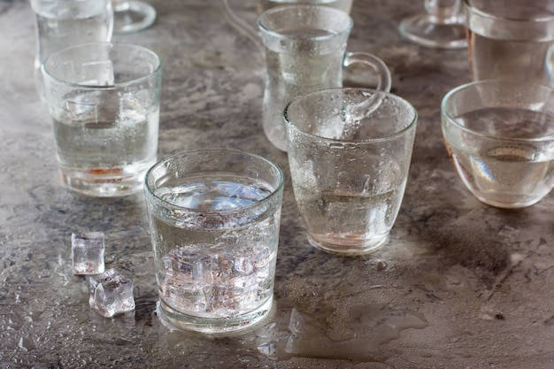 Muchos vasos de agua salpicados con gotas y trozos de hielo sobre una mesa gris